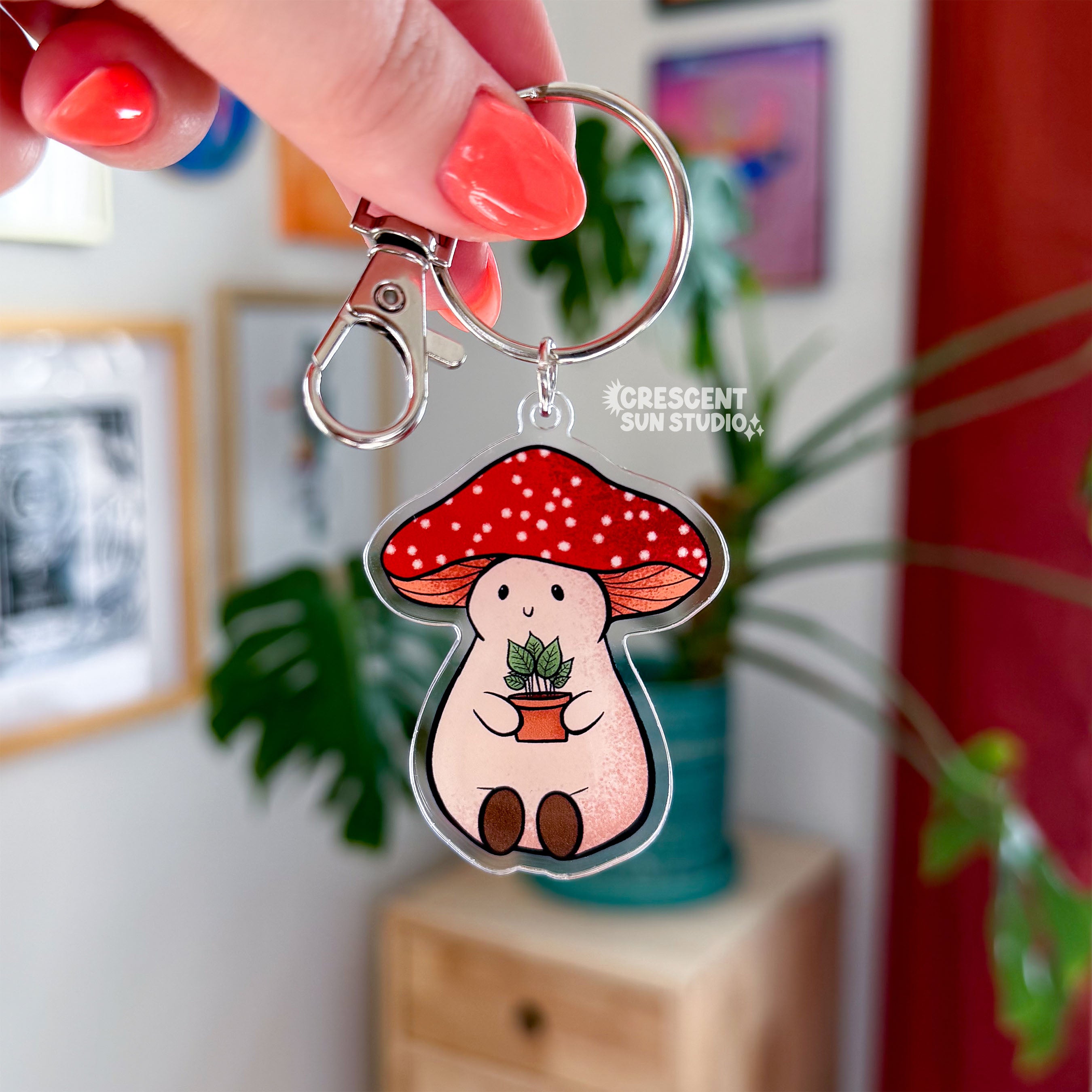 Stoney the Mushroom is a plump, smiling pinky-beige mushroom character with a red mushroom cap with white spots. Stoney is holding a tiny potted plant in his little mushroom arms. This acrylic keychain is topped with a silver lobster clasp and ring, which is being held by a woman's hand with coral painted fingernails. 

There is a vibrant green plant behind the keychain.