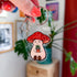 Stoney the Mushroom is a plump, smiling pinky-beige mushroom character with a red mushroom cap with white spots. Stoney is holding a tiny potted plant in his little mushroom arms. This acrylic keychain is topped with a silver lobster clasp and ring, which is being held by a woman's hand with coral painted fingernails. 

There is a vibrant green plant behind the keychain.