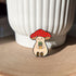 An enamel pin of a plump, smiling pinky-beige mushroom character with a red mushroom cap with white spots is holding a tiny potted plant in his little mushroom arms. The pin is depicted resting against a white pot.