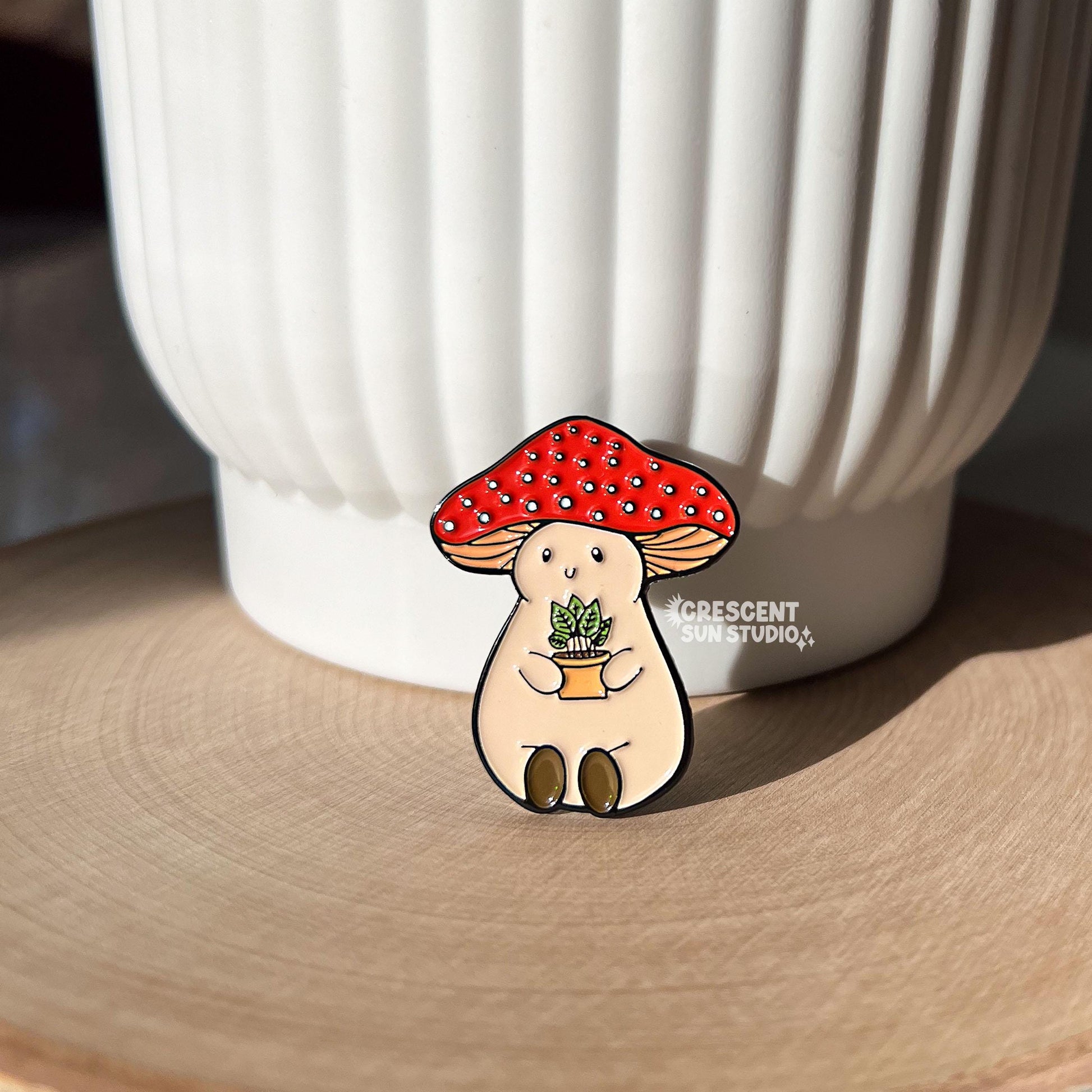 An enamel pin of a plump, smiling pinky-beige mushroom character with a red mushroom cap with white spots is holding a tiny potted plant in his little mushroom arms. The pin is depicted resting against a white pot.