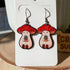 A close up image of our Stoney the Mushroom earrings. The earrings feature a plump, smiling pinky-beige mushroom character with a red mushroom cap with white spots. Stoney is holding a tiny potted plant in his little mushroom arms.