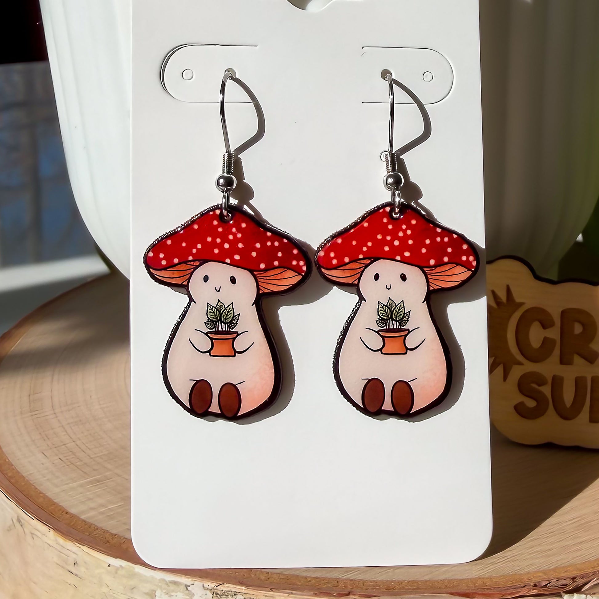 A close up image of our Stoney the Mushroom earrings. The earrings feature a plump, smiling pinky-beige mushroom character with a red mushroom cap with white spots. Stoney is holding a tiny potted plant in his little mushroom arms.