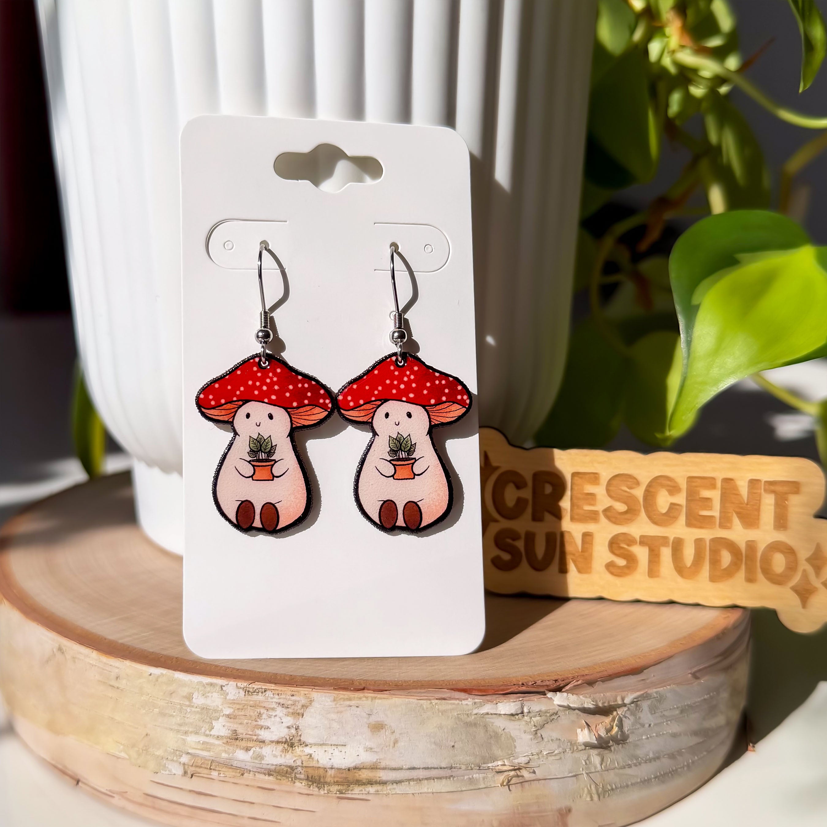 These earrings feature Stoney the Mushroom. He is a plump, smiling pinky-beige mushroom character with a red mushroom cap with white spots. Stoney is holding a tiny potted plant in his little mushroom arms.

In this image, the earrings are hanging on a white display card, which is propped up against a white plant pot.