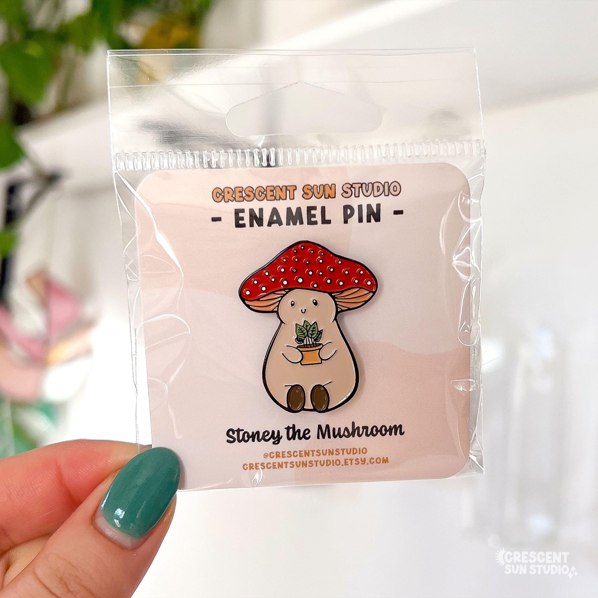 An enamel pin of a plump, smiling pinky-beige mushroom character with a red mushroom cap with white spots is holding a tiny potted plant in his little mushroom arms. The pin is displayed on a small card with the text "Crescent Sun Studio, enamel pin."