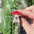 An enamel pin of a plump, smiling pinky-beige mushroom character with a red mushroom cap with white spots is holding a tiny potted plant in his little mushroom arms. A hand holds the pin close to the camera.