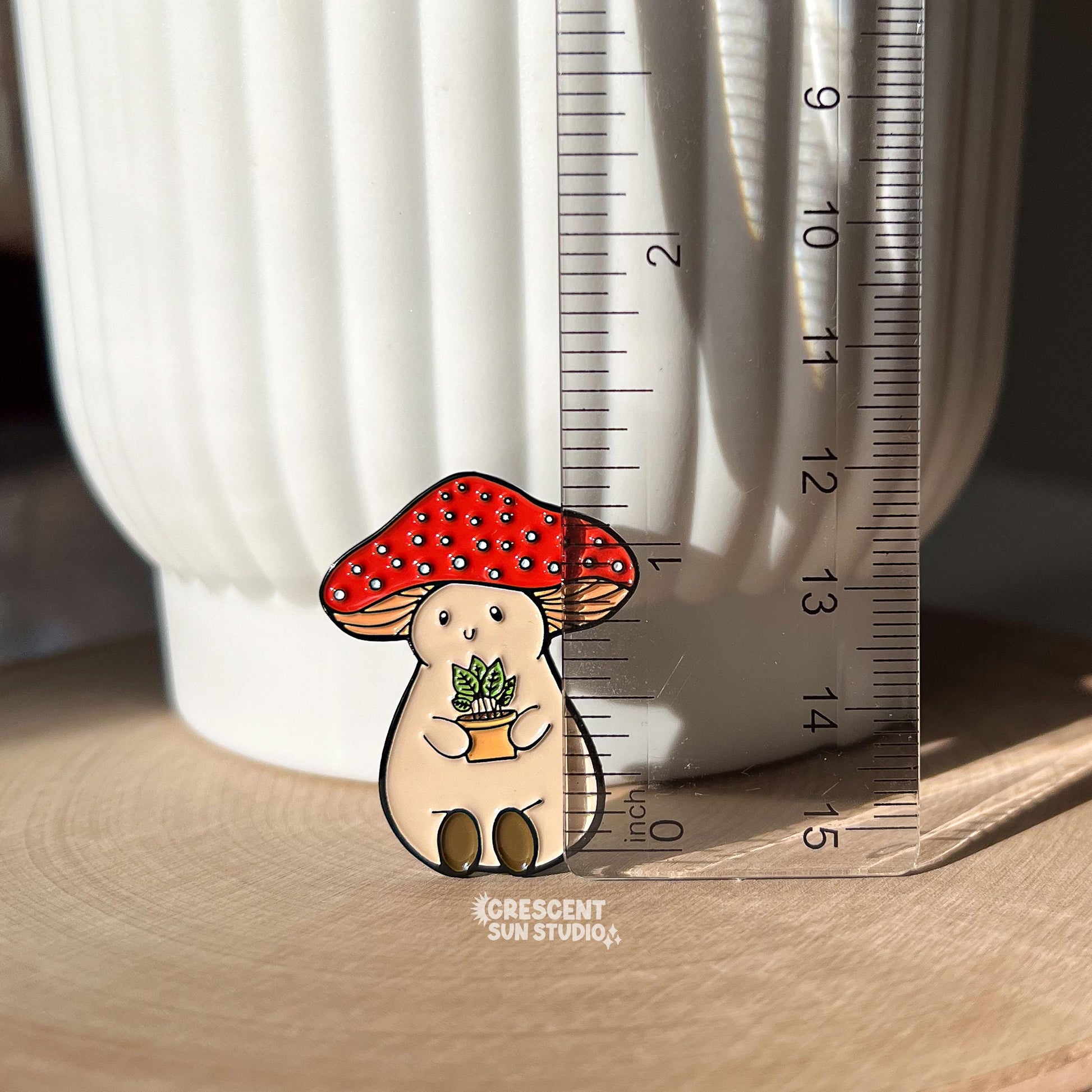 An enamel pin of a plump, smiling pinky-beige mushroom character with a red mushroom cap with white spots is holding a tiny potted plant in his little mushroom arms. This image has the pin next to a clear ruler, showing its size of 1.35 inches.