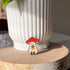 An enamel pin of a plump, smiling pinky-beige mushroom character with a red mushroom cap with white spots is holding a tiny potted plant in his little mushroom arms. The pin is leaning against a large white plant pot.