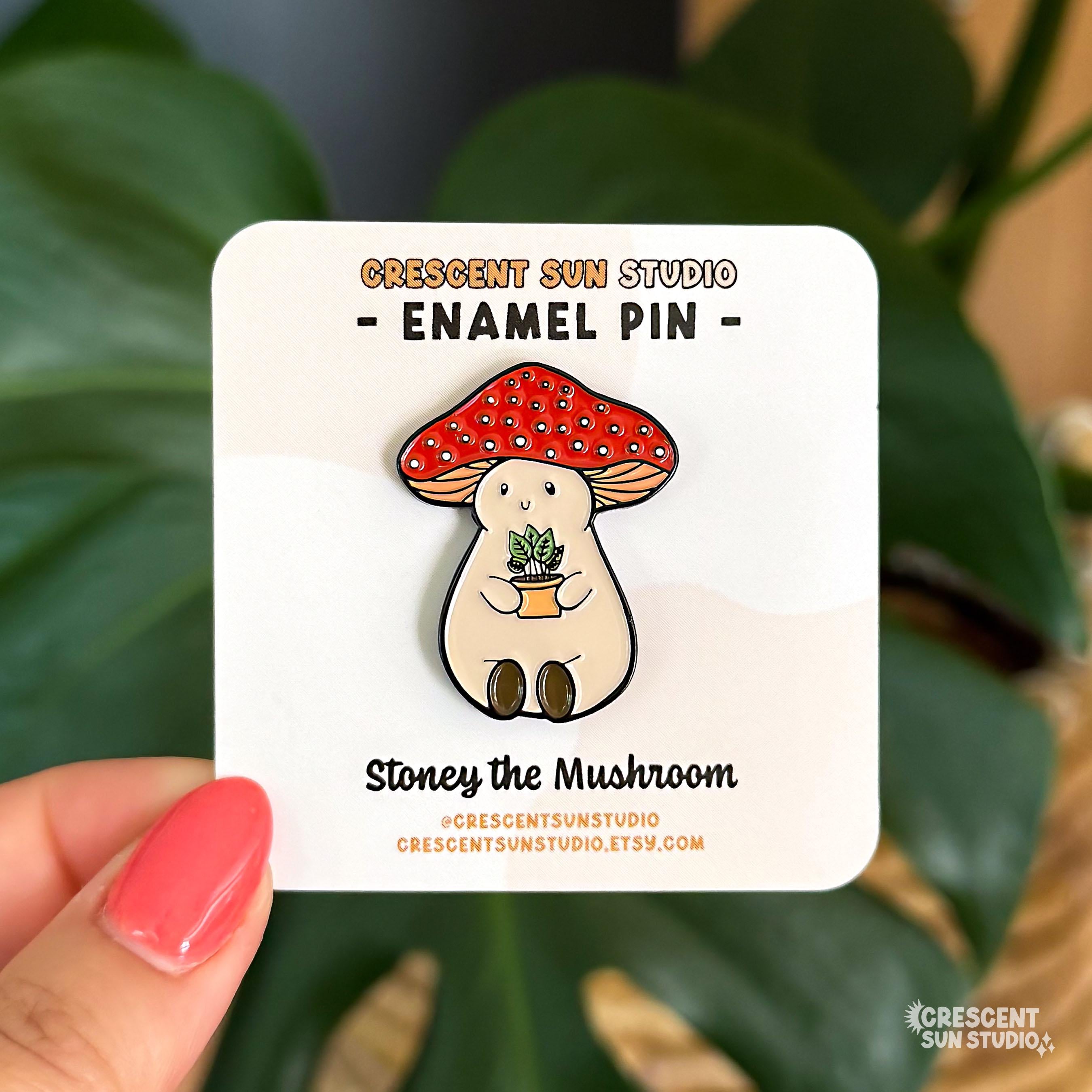 An enamel pin of a plump, smiling pinky-beige mushroom character with a red mushroom cap with white spots is holding a tiny potted plant in his little mushroom arms. The pin is displayed on a small card with the text "Crescent Sun Studio, enamel pin."