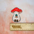 This image shows Stoney sitting above a ruler, showing the magnet's width of 2.4 inches. 

Stoney is a plump, smiling pinky-beige mushroom character with a red cap with white spots. Stoney is holding a tiny potted plant in his little mushroom arms. 