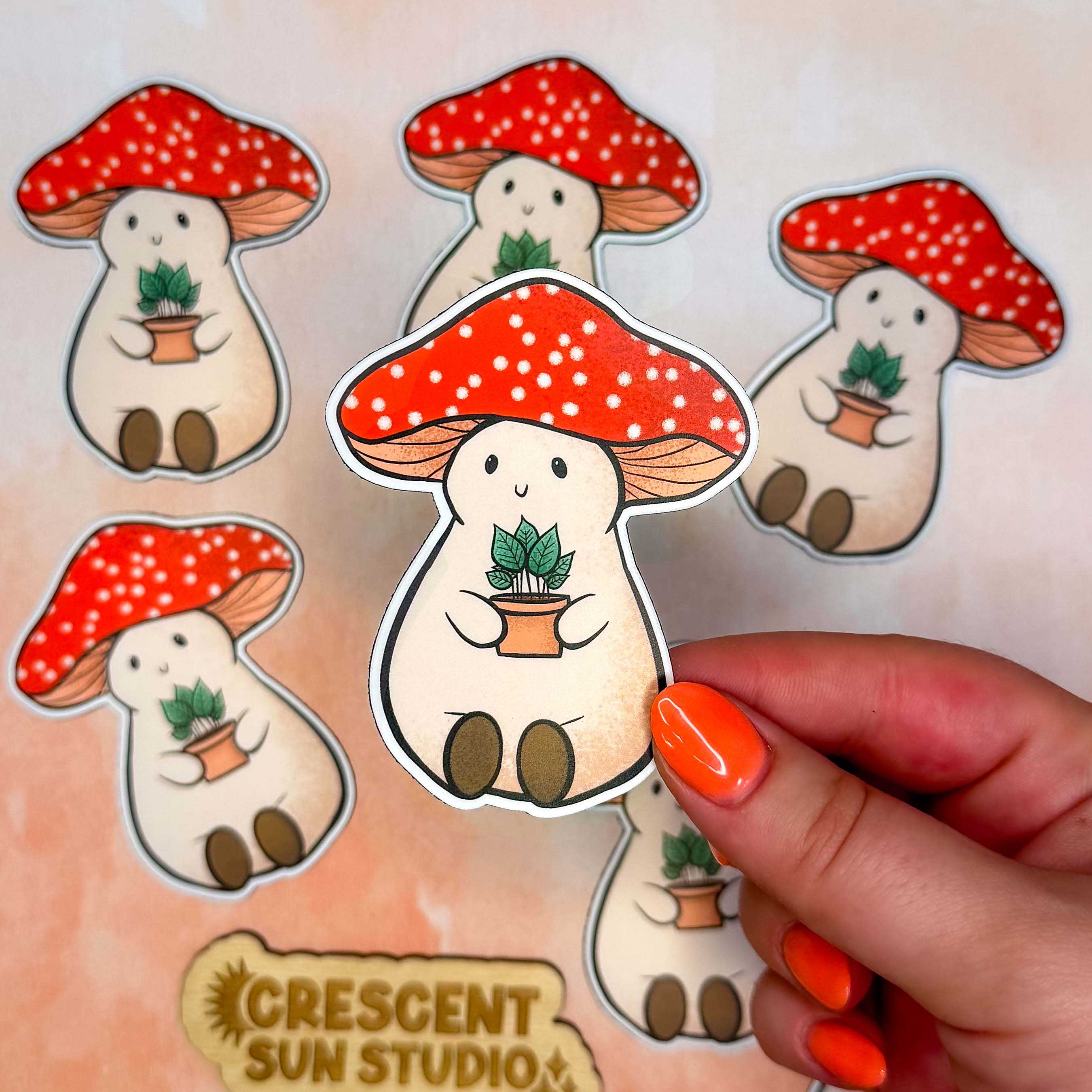 This image features several Stoney the Mushroom magnets, with one being held closer to the camera by a hand with coral orange painted fingernails. 

Stoney the Mushroom is a plump, smiling pinky-beige mushroom character with a red mushroom cap with white spots. Stoney is holding a tiny potted plant in his little mushroom arms. 