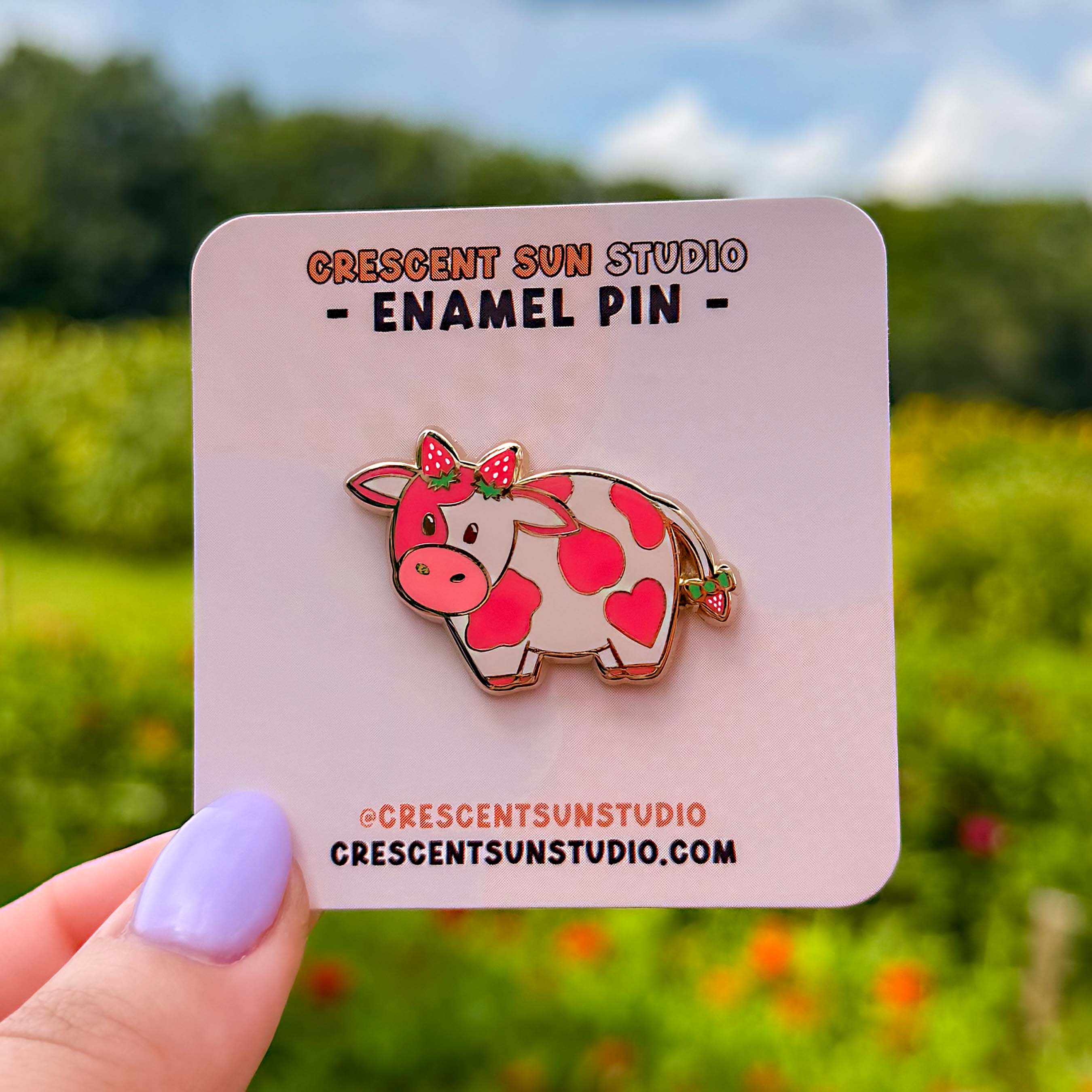 A small enamel pin of a pink cow with darker pink spots has two strawberries at the top of her head, as if they were horns, as well as a strawberry at the end of her tail. The pin is displayed on a light pink card.