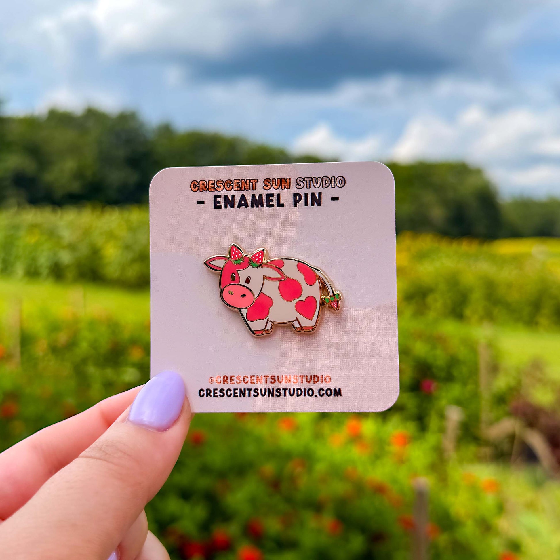 A small enamel pin of a pink cow with darker pink spots has two strawberries at the top of her head, as if they were horns, as well as a strawberry at the end of her tail. The pin is displayed on a light pink card.