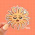 This sticker features a blissed out and glam sun with rays of light dancing around her. This personified sun has thin eyebrows, closed eyes with full lashes and eyeshadow, a nose ring, blushing cheeks, and glossy full lips.