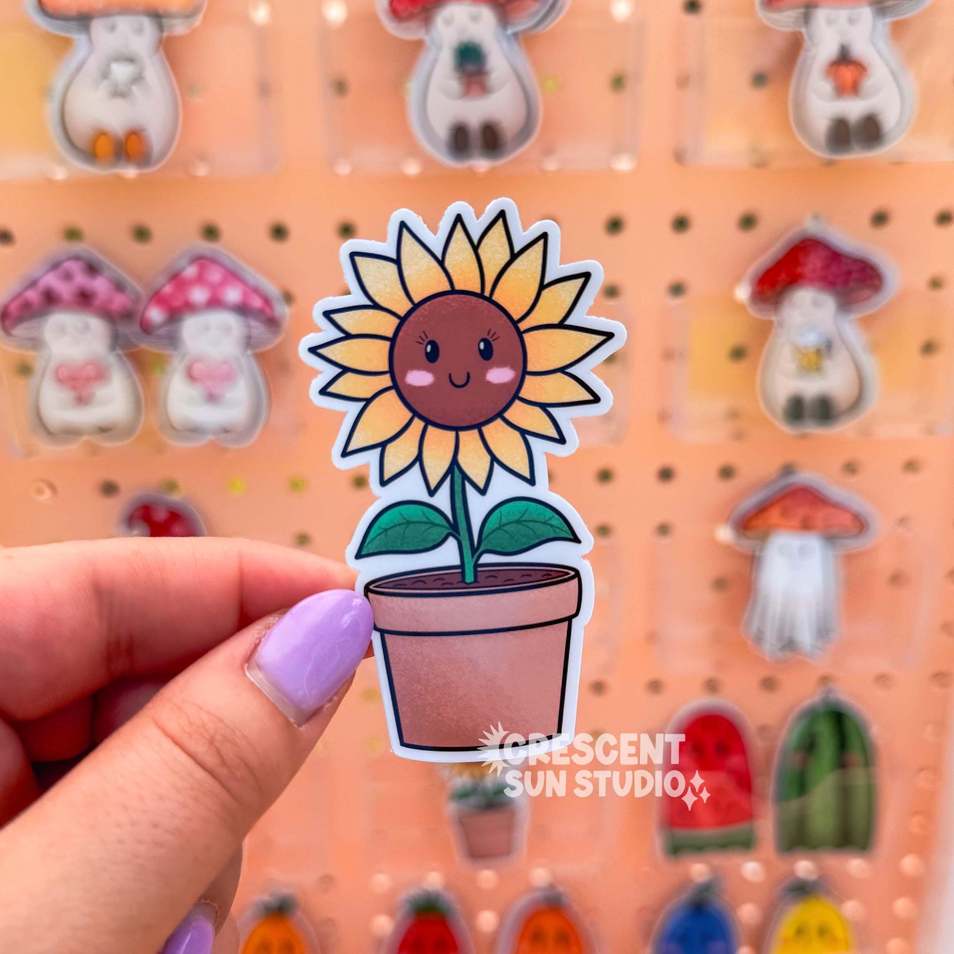 A smiling sunflower character with pink cheeks stands tall in a terracotta pot. The sunflower has yellow leaves and a warm brown face.

In this image, a hand holds the sticker up to the camera. In the background are rows of other stickers.