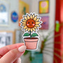 A smiling sunflower character with pink cheeks stands tall in a terracotta pot. The sunflower has yellow leaves and a warm brown face.