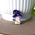 An enamel pin in the shape of a mushroom with an oversized mushroom cap that hooks to one side. The happy beige mushroom is holding a want with a crystal and moon on one end. His hat is purple with stars and spirals.