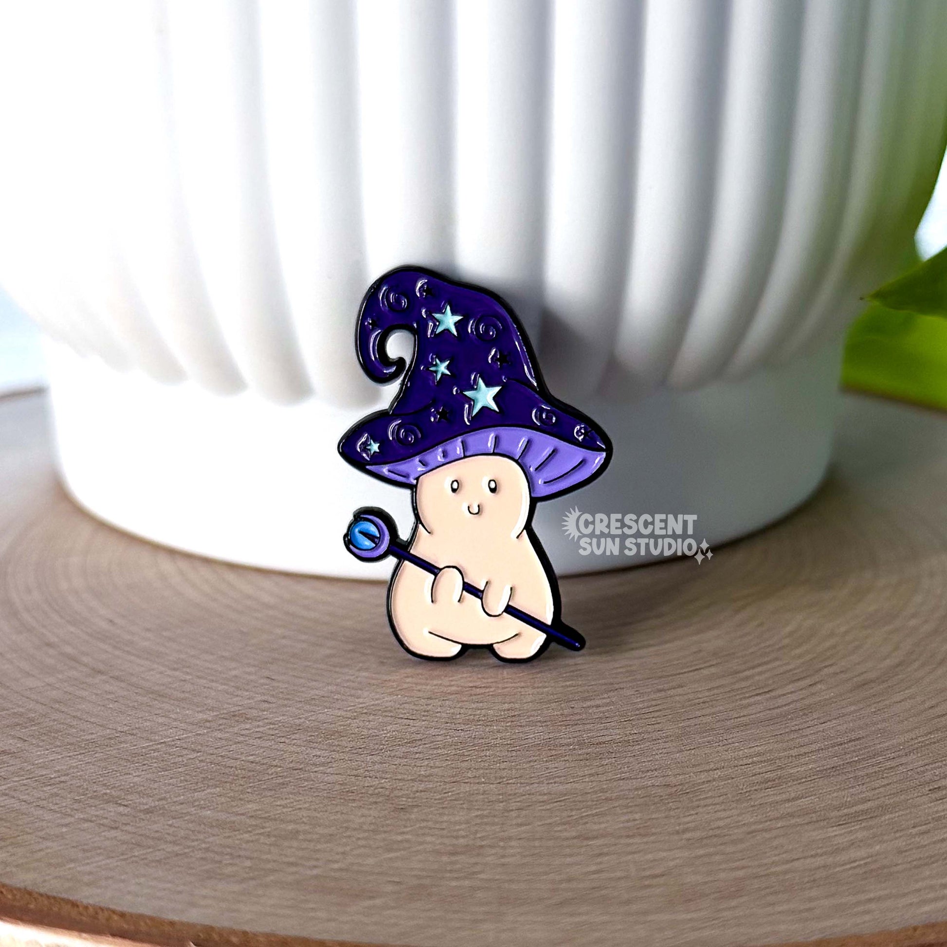 An enamel pin in the shape of a mushroom with an oversized mushroom cap that hooks to one side. The happy beige mushroom is holding a want with a crystal and moon on one end. His hat is purple with stars and spirals.