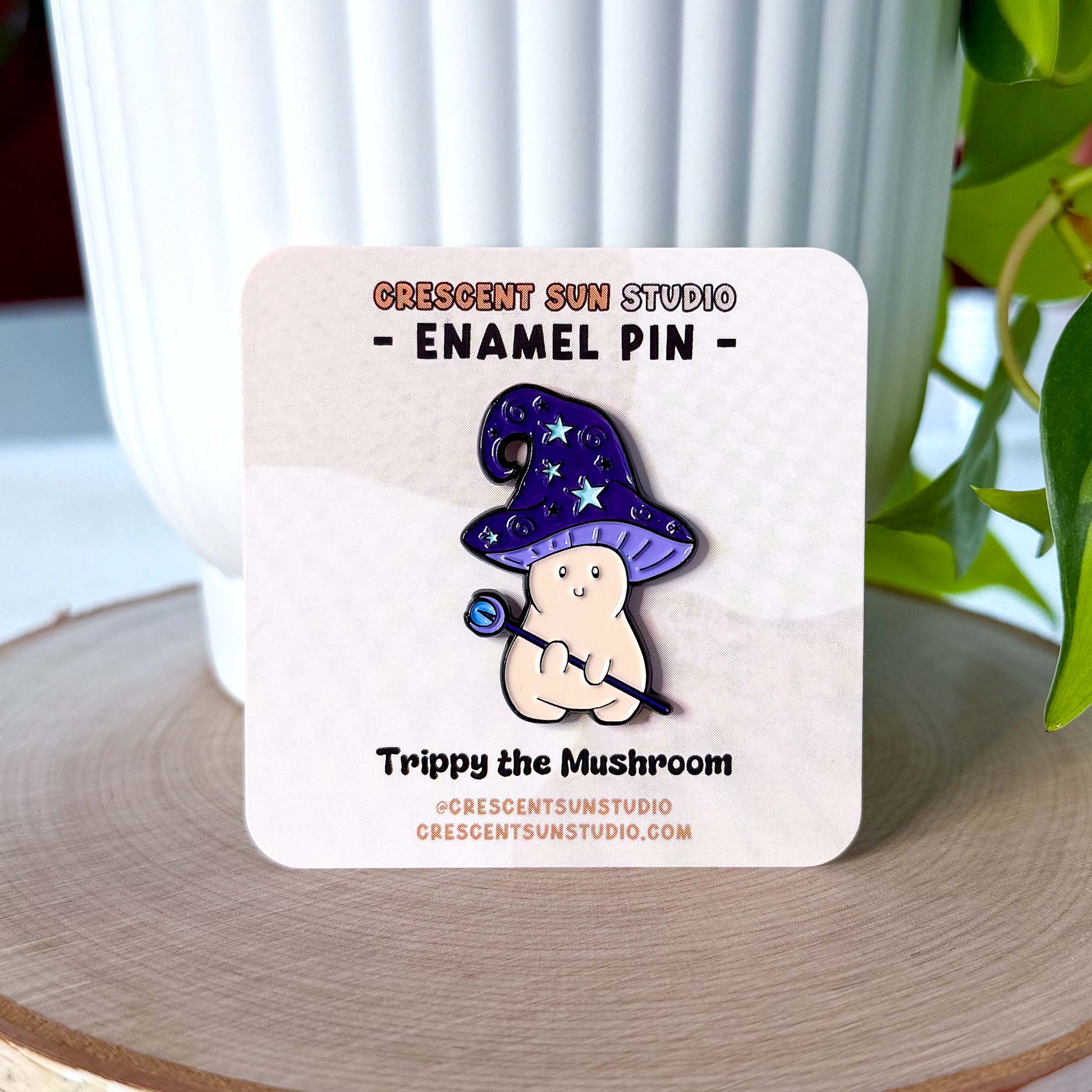 An enamel pin in the shape of a mushroom with an oversized mushroom cap that hooks to one side. The happy beige mushroom is holding a want with a crystal and moon on one end. His hat is purple with stars and spirals. In this image, the pin is displayed on a light colored card.