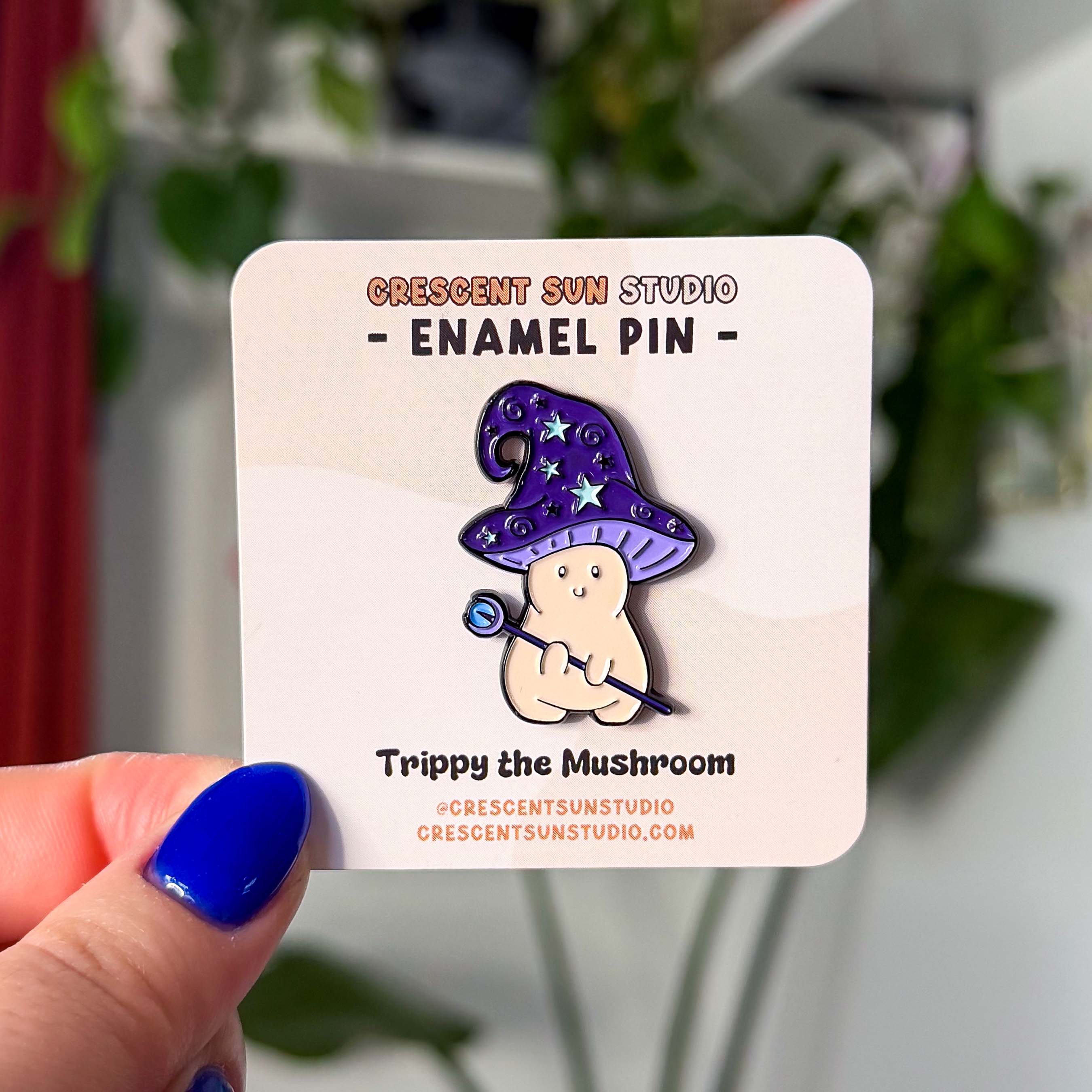 An enamel pin in the shape of a mushroom with an oversized mushroom cap that hooks to one side. The happy beige mushroom is holding a want with a crystal and moon on one end. His hat is purple with stars and spirals.