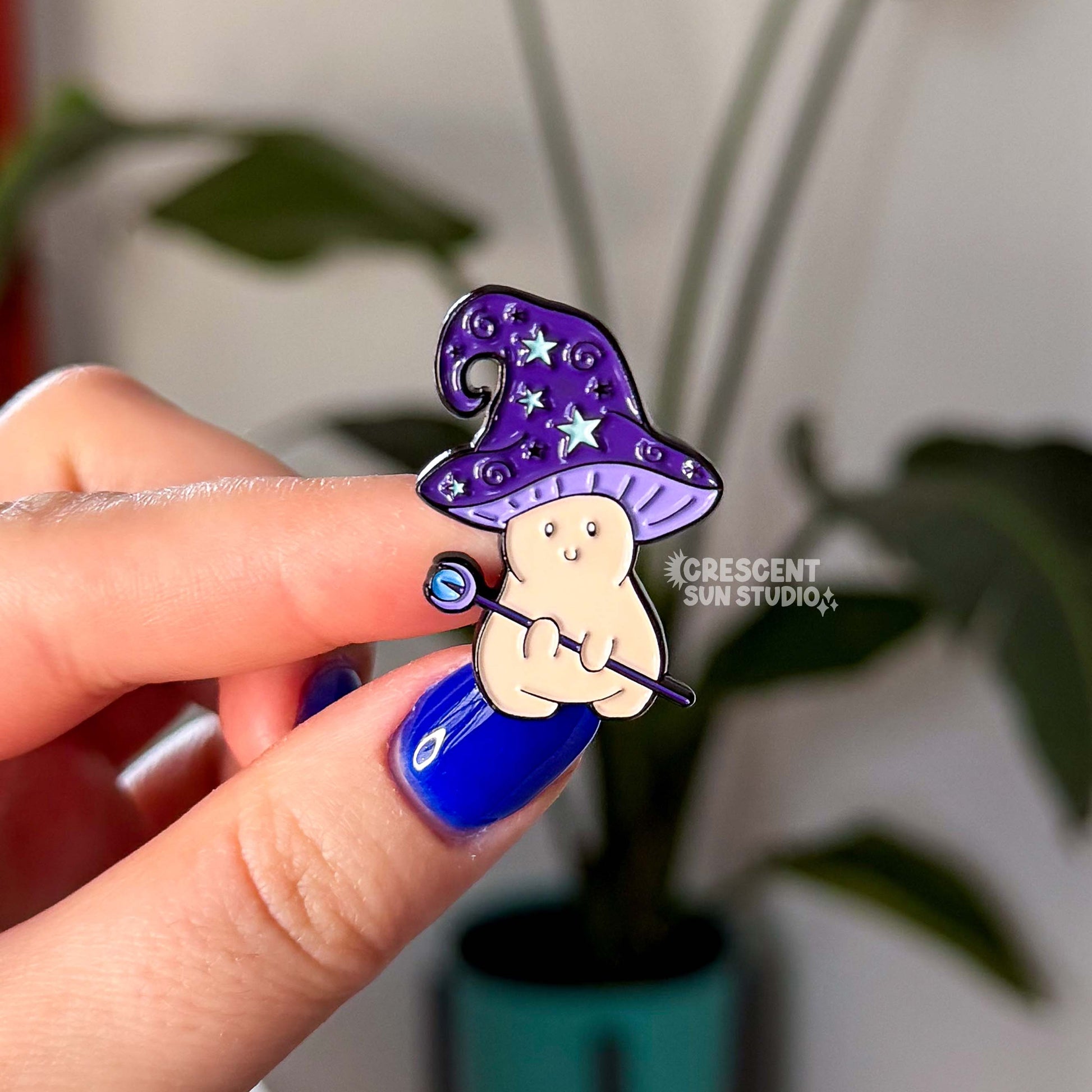 An enamel pin in the shape of a mushroom with an oversized mushroom cap that hooks to one side. The happy beige mushroom is holding a want with a crystal and moon on one end. His hat is purple with stars and spirals.