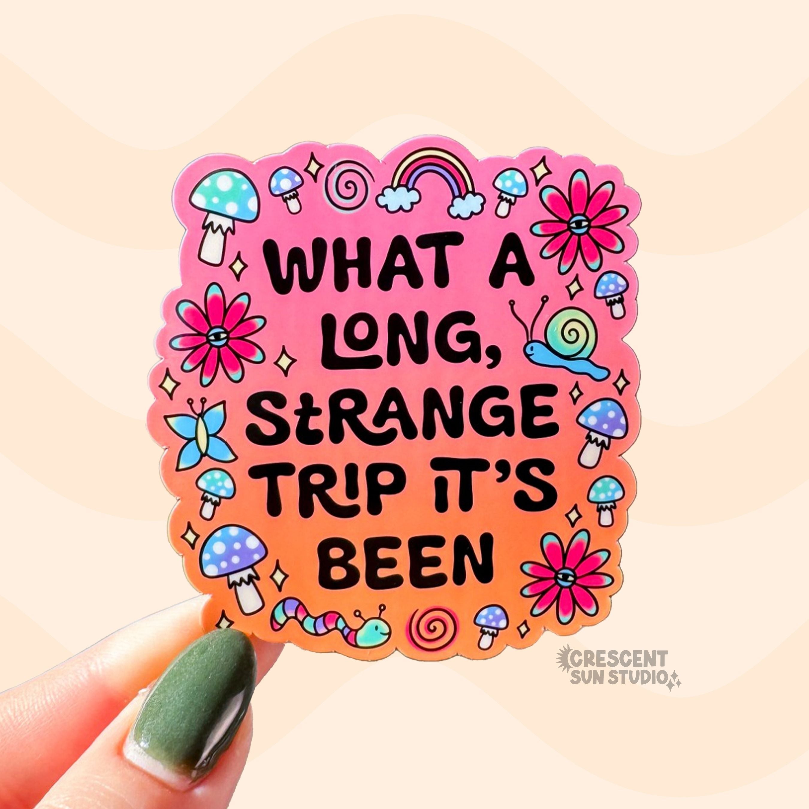 Die-cut sticker with a pink to orange gradient background reading 'What a Long Strange Trip It's Been' in black lettering surrounded by illustrated mushrooms, flowers with eyes, snails, a butterfly, and a worm.