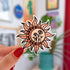 A pale orange die cut sticker in the shape of the sun with dancing rays. Inside the circle of the sun is a darker orange crescent and black doodles of wildflowers and vines.