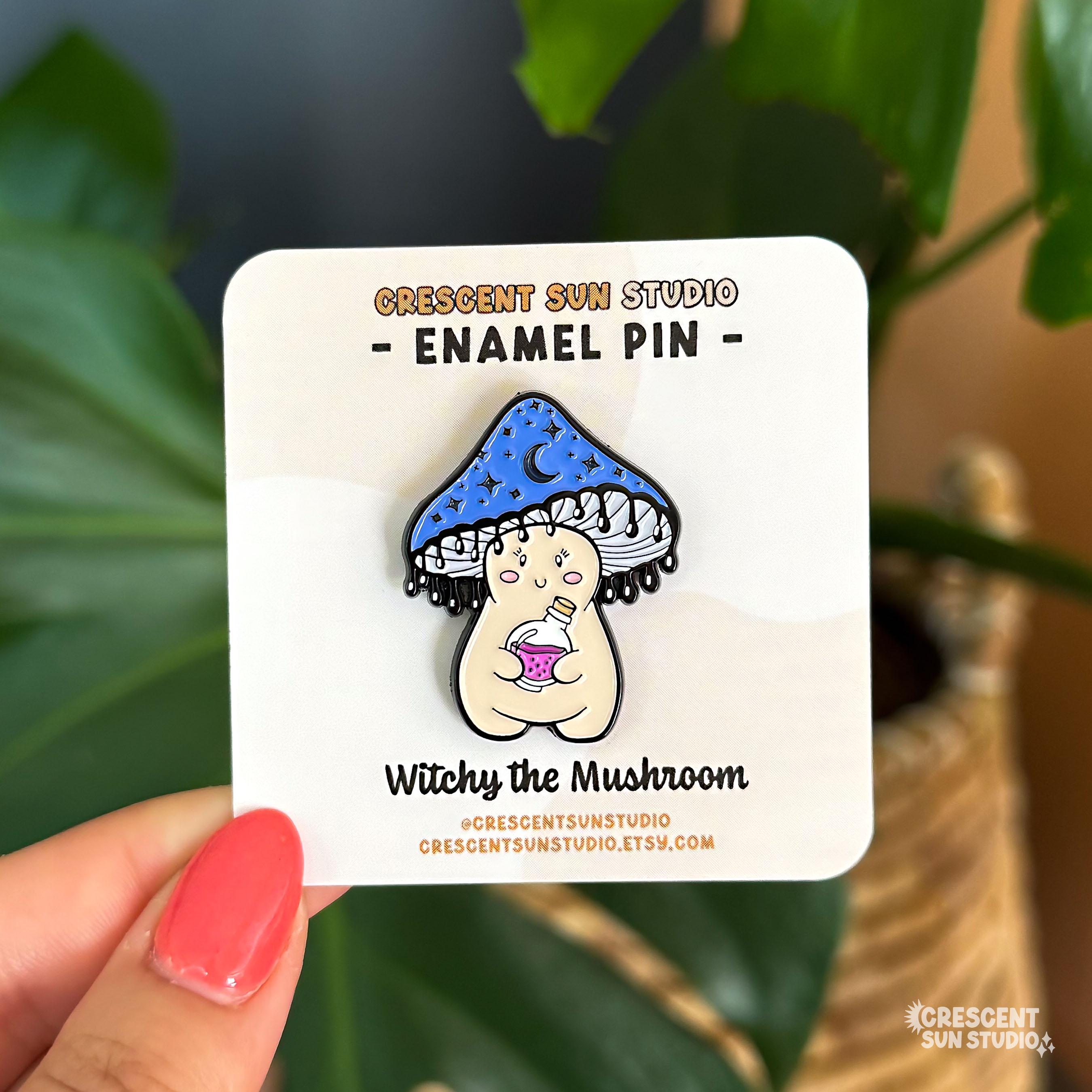 A pin in the shape of a happy little mushroom character. Her mushroom cap is shaded blue-black with dark moons and stars on it. The bottom of the cap has little drips, resembling ink. She is holding a potion bottle with pink liquid inside.