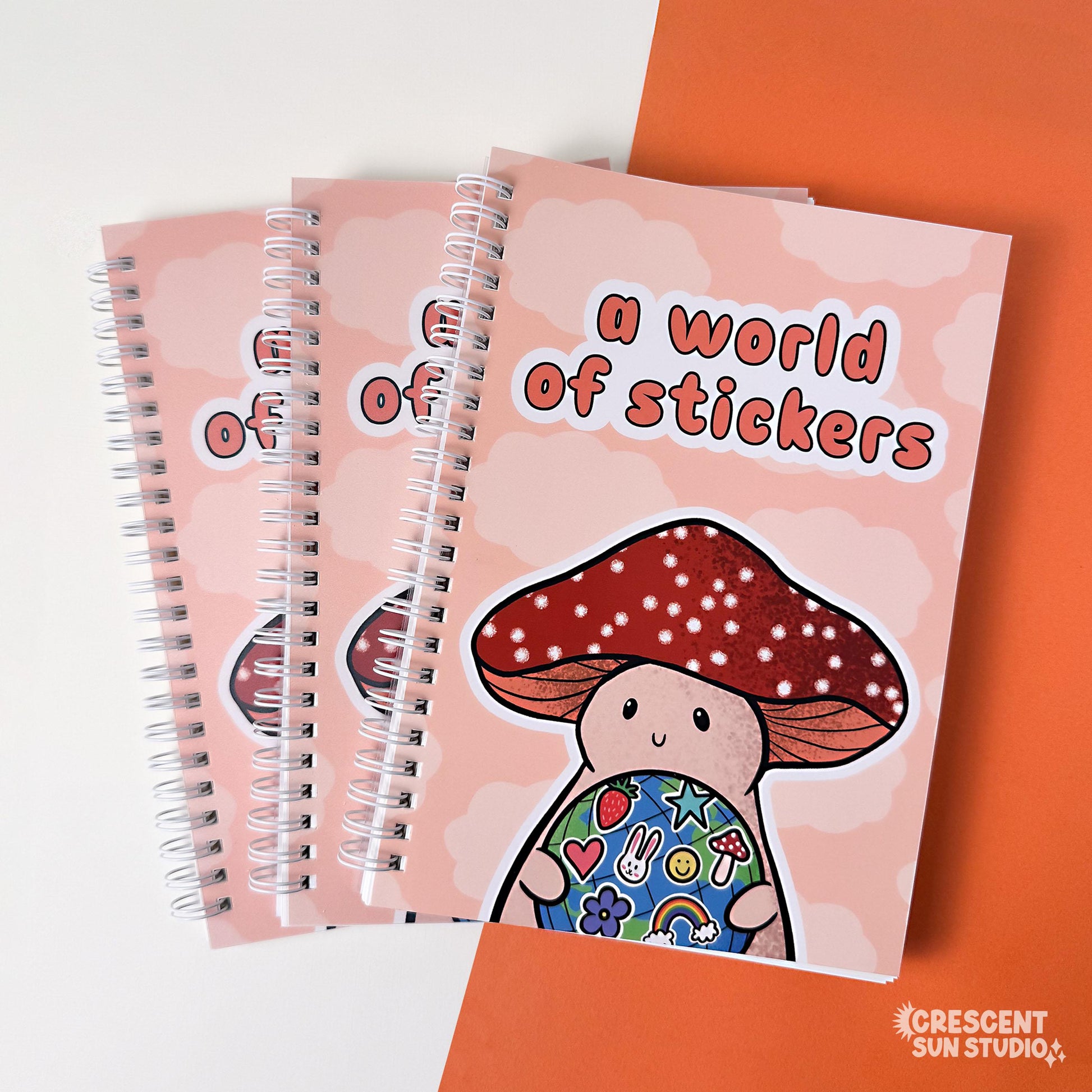 Spiral bound book with a background of peachy clouds. Coral text reads "a world of stickers." Below that is a happy mushroom holding a globe covered in stickers.